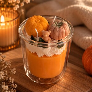 Könnte beinhalten: Ein transparenter Glasbehälter gefüllt mit orangefarbenem Wachs, garniert mit weißem Wachs in Schlagsahneoptik, einem leuchtend orangefarbenen Kürbis, einem blassrosa Kürbis, einer kleinen beigen Bärenfigur und dunkelgrünen Blattakzenten. Die Kerze hat einen Docht.