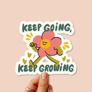 Peut inclure: Un autocollant découpé représentant une fleur rose avec un visage jaune et des yeux fermés, marchant avec des bras et des jambes. Le texte "KEEP GOING, KEEP GROWING" est imprimé en vert foncé au-dessus et en dessous, avec des éclats jaunes.