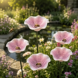 Puede incluir: Cinco estacas decorativas de jardín con flores de vidrio de color rosa pálido en forma de cuenco y textura agrietada. Cada flor está montada sobre un tallo de metal oscuro y fino, con varias abejas posadas dentro de los pétalos.