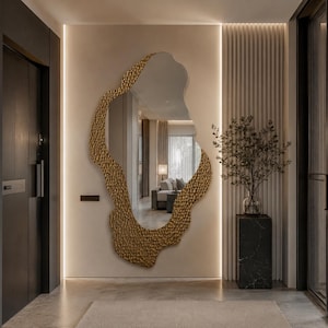 May include: A large, irregular, organic-shaped wall mirror featuring a textured, metallic gold frame with a hammered finish. The mirror is mounted on a light-colored wall, reflecting a living space with a sofa and window treatments.