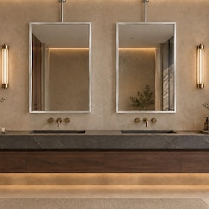 May include: A double vanity bathroom setup featuring a long dark grey stone countertop with two integrated sinks, mounted above a floating wooden cabinet. Two rectangular mirrors with silver frames hang above, flanked by vertical wall sconces.