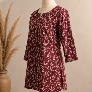 May include: A maroon tunic featuring a cream-coloured botanical leaf print, three-quarter length sleeves, a round neckline with a small front slit, and side hem vents. The garment is displayed on a mannequin, showcasing a straight-cut silhouette.