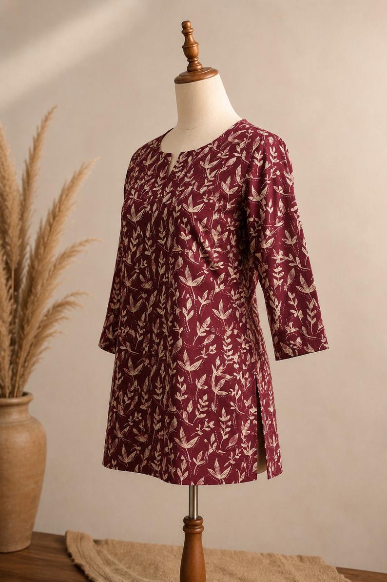 May include: A maroon tunic featuring a cream-coloured botanical leaf print, three-quarter length sleeves, a round neckline with a small front slit, and side hem vents. The garment is displayed on a mannequin, showcasing a straight-cut silhouette.