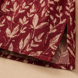 May include: A deep red garment featuring a repeating cream-coloured botanical leaf pattern. The fabric displays a straight hemline with a vertical side slit, showcasing the detailed printed foliage design across the entire visible surface area.