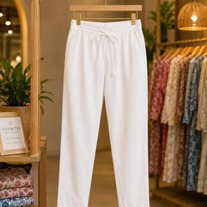 May include: A pair of solid white trousers featuring an elasticized waistband with a matching white drawstring tie. The pants have a straight-leg silhouette with small vertical slits at the bottom of each hem, hanging on a wooden hanger.