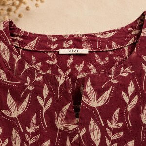 May include: A deep maroon shirt featuring a cream-coloured botanical leaf pattern throughout. The garment has a round neckline with a central vertical slit opening and a white interior label with the text VIVE printed in black.