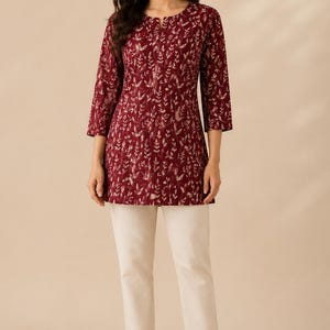 May include: A maroon tunic featuring a white botanical leaf print, a round neckline with a small slit, and three-quarter length sleeves, paired with straight-leg cream-coloured trousers and tan heeled sandals with a T-strap design.