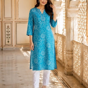 Puede incluir: Una túnica de color turquesa brillante hasta la rodilla con un estampado floral de puntos blancos, escote con abertura y mangas tres cuartos, combinada con pantalones rectos blancos lisos y sandalias abiertas de color claro.