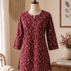 May include: A deep burgundy tunic featuring a repeating cream-coloured botanical leaf pattern. The garment has three-quarter length sleeves, a straight hemline with side slits, and a small notched neckline, displayed on a dress form mannequin.