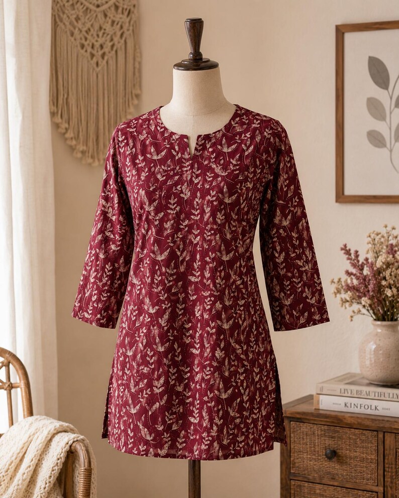 May include: A deep burgundy tunic featuring a repeating cream-coloured botanical leaf pattern. The garment has three-quarter length sleeves, a straight hemline with side slits, and a small notched neckline, displayed on a dress form mannequin.