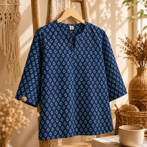 May include: A dark blue tunic with a white repeating geometric floral pattern, featuring three-quarter length sleeves and a notched V-neckline. The garment has a straight hem and a small white tag at the inner collar labeled with the letter M.
