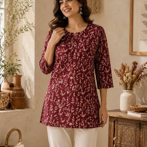 May include: A maroon tunic featuring a white botanical leaf print, three-quarter length sleeves, and a small notch neckline. The garment is paired with plain white trousers, creating a simple and clean look with a consistent pattern throughout.