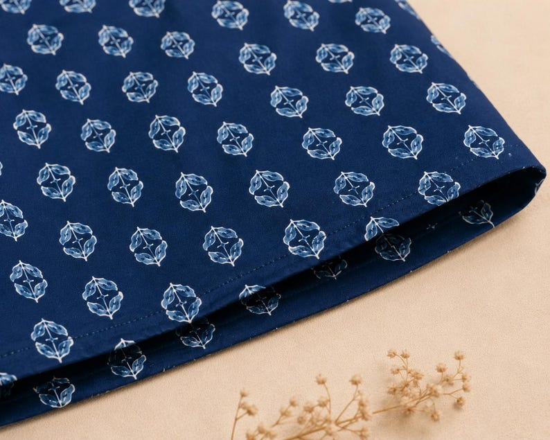 May include: A dark blue fabric featuring a repeating white botanical pattern of small, stylized leaves arranged in a symmetrical diamond-like formation. The material has a visible stitched hem along the bottom edge, showcasing a clean finish.