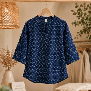 May include: A dark blue blouse with a repeating white geometric pattern, featuring three-quarter length sleeves, a split neckline, and a straight hem. The garment is displayed on a wooden hanger and includes a small white tag at the collar.
