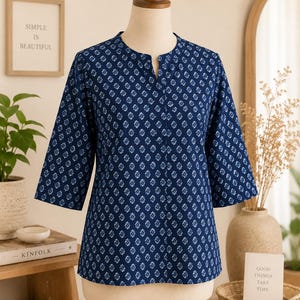 May include: A dark blue blouse with a repeating white geometric pattern, featuring three-quarter length sleeves, a mandarin collar, and a small V-shaped slit at the neckline. The garment is displayed on a mannequin, showcasing a straight hemline.