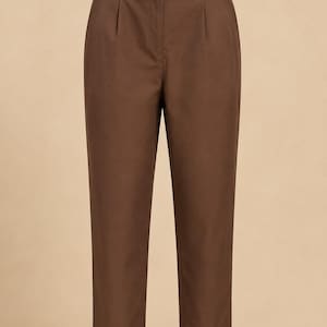 Puede incluir: Pantalones de color marrón sólido con diseño de cintura alta, pretina elástica, un pequeño lazo decorativo en el centro de la parte delantera y pliegues sutiles debajo de la cintura. Estos pantalones tienen un corte cónico y acabado limpio.