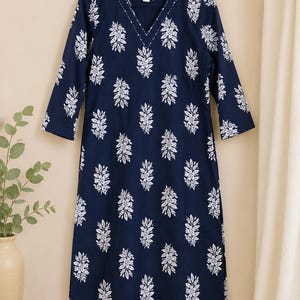 Può includere: Abito blu navy con motivo floreale bianco. L'abito ha uno scollo a V e maniche a tre quarti. Il motivo floreale è ripetuto sul tessuto. L'abito è appeso a una gruccia di legno.