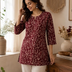 May include: A maroon tunic featuring a repeating cream-coloured botanical leaf pattern, three-quarter length sleeves, and a small notch neckline. The garment is paired with plain off-white trousers, creating a coordinated ensemble with a consistent print.