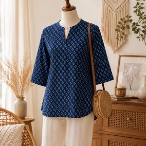 May include: A dark blue blouse with a white geometric floral pattern, featuring a split V-neckline and three-quarter length sleeves. The blouse is paired with plain off-white trousers and a round, woven straw crossbody bag with a brown strap.