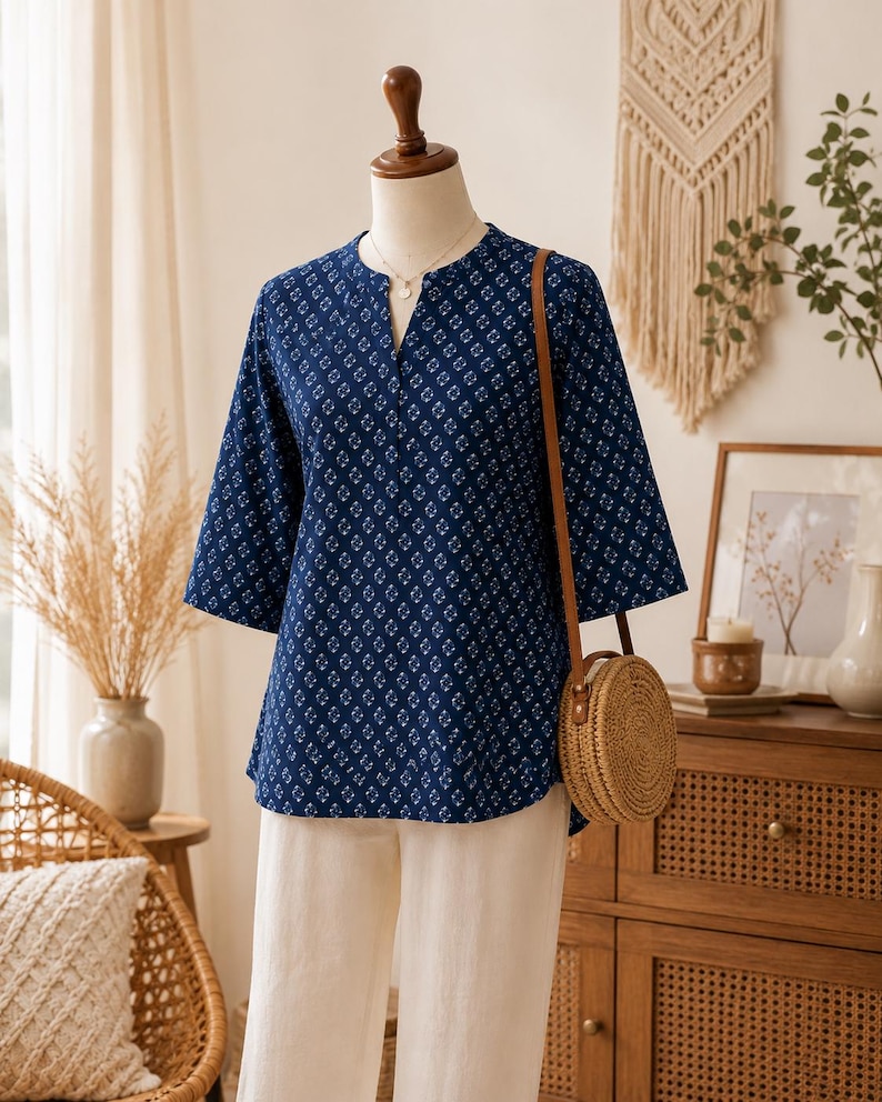 May include: A dark blue blouse with a white geometric floral pattern, featuring a split V-neckline and three-quarter length sleeves. The blouse is paired with plain off-white trousers and a round, woven straw crossbody bag with a brown strap.