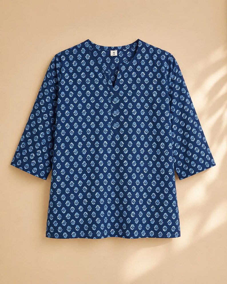 May include: A dark blue tunic with three-quarter length sleeves and a split V-neckline. The garment features an all-over repeating light blue floral block print pattern. A small white tag with the letter M is visible at the inner neckline.