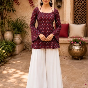 May include: A maroon long-sleeved tunic featuring a small repeating floral pattern in blue and white, paired with solid white wide-leg trousers. The tunic has a square neckline with thin straps and flared sleeves that extend past the wrists.