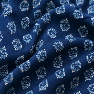 May include: A deep blue fabric featuring a repeating pattern of white and light blue stylized floral motifs. The material is draped to show soft folds, highlighting the consistent geometric arrangement of the small, symmetrical botanical shapes.