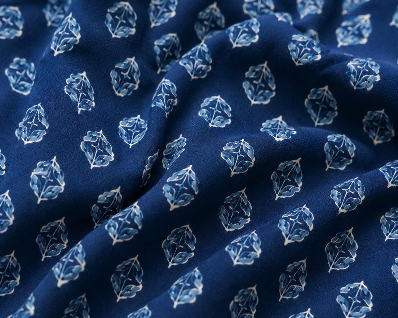 May include: A deep blue fabric featuring a repeating pattern of white and light blue stylized floral motifs. The material is draped to show soft folds, highlighting the consistent geometric arrangement of the small, symmetrical botanical shapes.