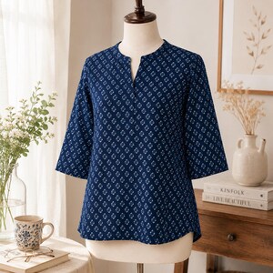 May include: A dark blue blouse featuring a repeating white geometric diamond pattern, three-quarter length sleeves, and a split neckline. The garment is displayed on a mannequin, showcasing a straight hemline and a structured fit throughout the torso.