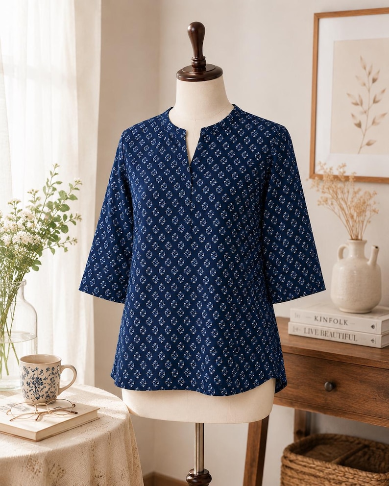 May include: A dark blue blouse featuring a repeating white geometric diamond pattern, three-quarter length sleeves, and a split neckline. The garment is displayed on a mannequin, showcasing a straight hemline and a structured fit throughout the torso.