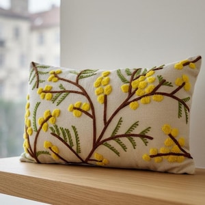 Puede incluir: Cojín decorativo rectangular con bordado de flores de mimosa amarillas y ramas marrones sobre fondo beige. El cojín está colocado sobre una superficie de madera, mostrando el diseño floral.
