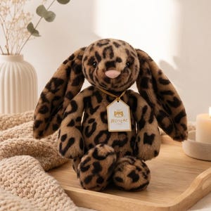 Puede incluir: Un peluche de conejito con estampado de leopardo. El juguete tiene orejas largas, una nariz rosa y una etiqueta que dice "Jellycat". El conejito está sentado en una bandeja de madera.