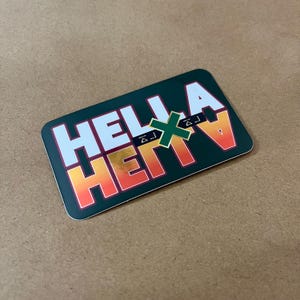 May include: A rectangular sticker with a dark green background featuring the white text HELLA and a gradient orange to yellow text HEAVY. A small green X symbol is positioned in the center between the two words, with small geometric accents.