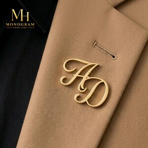 May include: A gold-toned metal brooch featuring the intertwined letters AD in a cursive script font, pinned to a tan blazer lapel. The text MH MONOGRAM HAND CARE is visible in the upper left corner against a black fabric background.