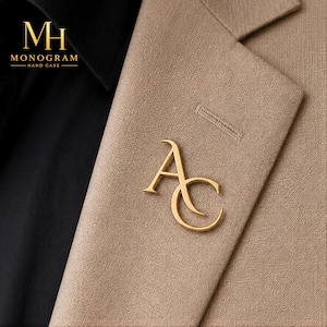 Könnte beinhalten: Ein goldfarbener Monogramm-Anstecker mit den Buchstaben "AC" ist an einem beigefarbenen Blazer befestigt. Der Anstecker befindet sich auf dem Revers des Blazers. In der oberen linken Ecke des Bildes befindet sich das Logo "MH MONOGRAM HAND CARE".
