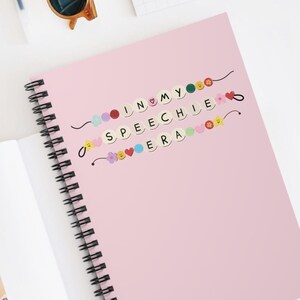 Könnte beinhalten: Ein spiralgebundenes Notizbuch mit einem hellrosa Einband, das eine Grafik aus bunten Perlen zeigt, die den Text IN MY SPEECHIE ERA bilden, akzentuiert durch kleine Blumen- und Herzformen. Eine goldene Metallklemme ist an der offenen Seite befestigt.