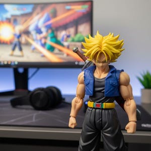 Puede incluir: Figura de acción detallada de un personaje con pelo amarillo puntiagudo, que lleva un chaleco azul y pantalones negros, con una espada en la espalda. La figura está delante de un monitor de ordenador que muestra una escena de videojuego. También se ven una pequeña planta y unos auriculares.
