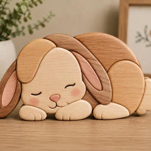 May include: A wooden bunny figurine in shades of light and dark brown, with pink accents on the ears and cheeks. The bunny is in a sleeping position, with its eyes closed and paws tucked in. The figurine is made of layered wood.