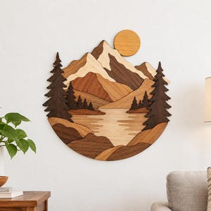 May include: A circular wall art piece crafted from layered wood in various natural brown tones. The design depicts a mountain range, a central lake, and evergreen trees on both sides, with a circular sun positioned in the upper right.