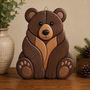 May include: A handcrafted wooden bear wall hanging. The bear is made of various shades of brown wood, with a lighter brown belly and paw pads. It has a black nose and eyes, and a metal hanger at the top. The bear is sitting.