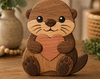 Op de afbeelding: Houten otterbeeldje met een hart. De otter is gemaakt van verschillende houttinten, met een lichtbruin gezicht, een donkerbruin lichaam en een rozerood hart. Het beeldje heeft een glimlachende uitdrukking en zit rechtop.