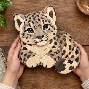 Puede incluir: Una pieza de arte mural de madera hecha a mano con forma de cachorro de leopardo de las nieves. El diseño presenta intrincados patrones de vetas de madera en tonos crema, bronceado y marrón oscuro, formando el pelaje manchado y las orejas.