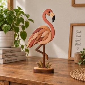 Könnte beinhalten: Eine dekorative Flamingo-Figur aus verschiedenen Naturholztönen mit geschwungenem Hals, einem schwarz gespitzten Schnabel und geschichteten Flügelsegmenten. Der Vogel steht auf einem Bein auf einem kleinen, ovalen Holzsockel.