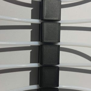 May include: A black 3D-printed cable management organizer featuring a vertical column of six rectangular blocks. Five white flexible tubes are threaded horizontally through the gaps between the blocks, creating a structured grid for cable routing.