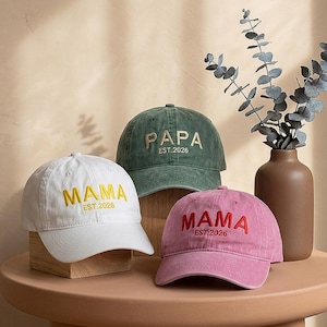 Puede incluir: Tres gorras de béisbol en blanco, verde oscuro y rosa empolvado. La gorra blanca tiene texto bordado en amarillo que dice MAMA EST.2026. La gorra verde tiene texto en beige que dice PAPA EST.2026. La gorra rosa tiene texto en rojo MAMA EST.2026.