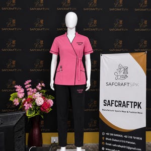 May include: A pink wrap-style scrub top with black trim and a side tie, featuring the text CUSTOM TEXT on the chest, paired with black scrub pants that also display the text CUSTOM TEXT on the left thigh, displayed on a white mannequin.