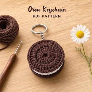 May include: A crocheted Oreo keychain with a silver keyring. The keychain is brown with a white cream filling. The image also includes a ball of brown yarn, a crochet hook, and a daisy. Text on the image reads "Oreo Keychain PDF Pattern."