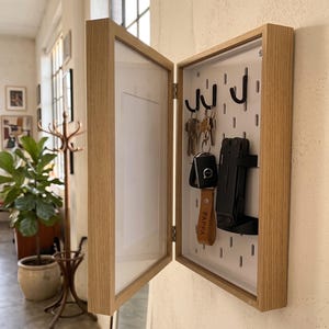 Pode incluir: Um armário de chaves de madeira clara montado na parede com porta articulada. O interior possui um painel perfurado branco com ganchos de metal preto que seguram várias chaves, um porta-chaves de carro e uma ferramenta multifunções preta.