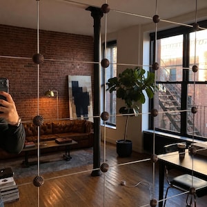 May include: A room divider featuring a grid of thin vertical and horizontal metal wires connected by small, round, dark brown decorative knobs. The divider is positioned in a room with a brick wall, a leather sofa, and a large potted plant.