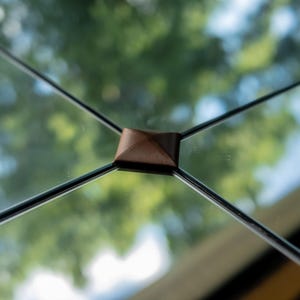 Op de afbeelding: Een vierkant bruin lederen verbindingsstuk gecentreerd op het snijpunt van vier dunne zwarte metalen staven. Het lederen onderdeel heeft een piramidevormig bovenoppervlak en dient als structurele verbinding voor de metalen frame-elementen.